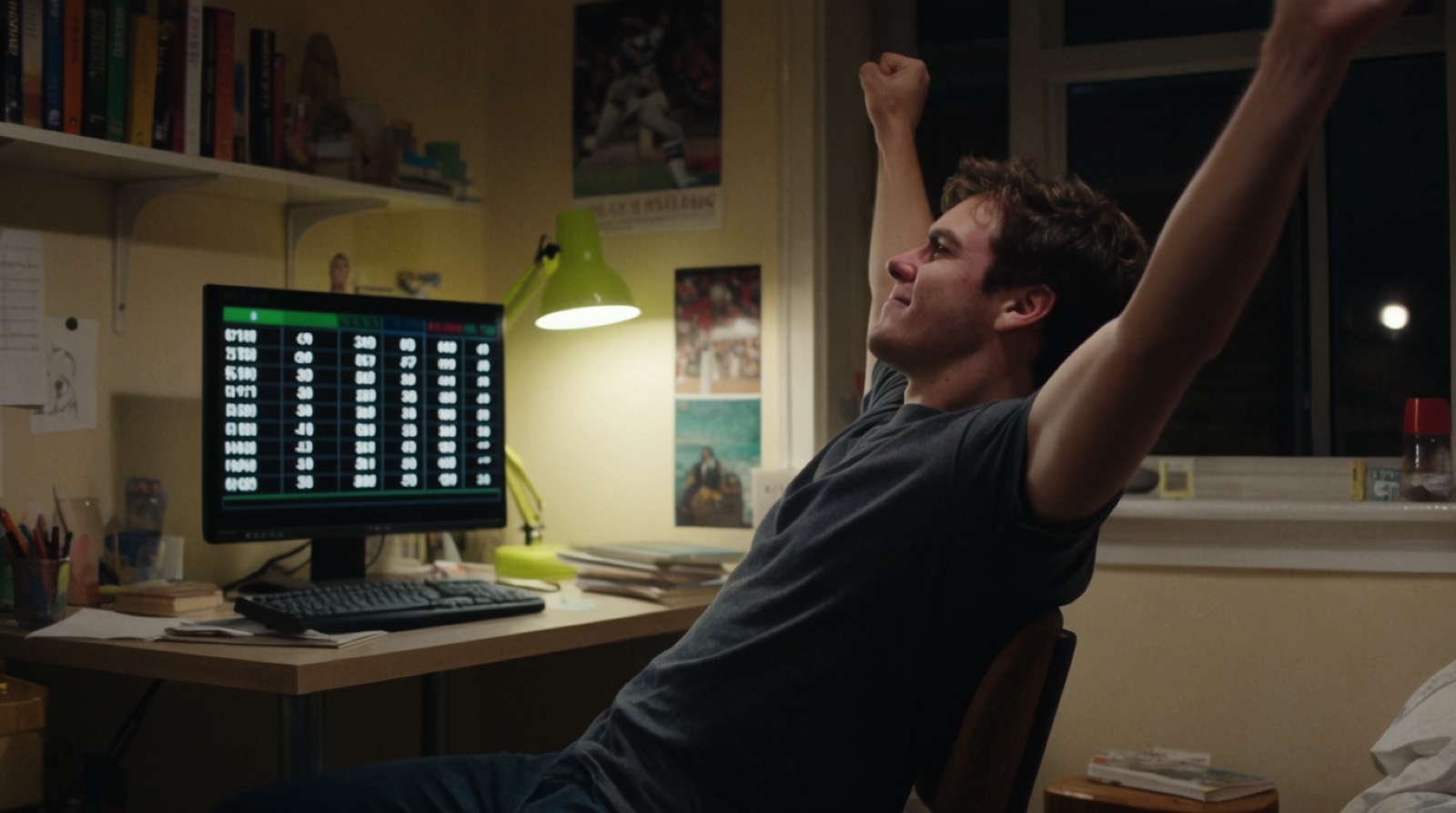 A man leaning back with both arms raised at a desk in a small room at night, a scoreboard glowing on the monitor, a lime desk lamp on the wall behind