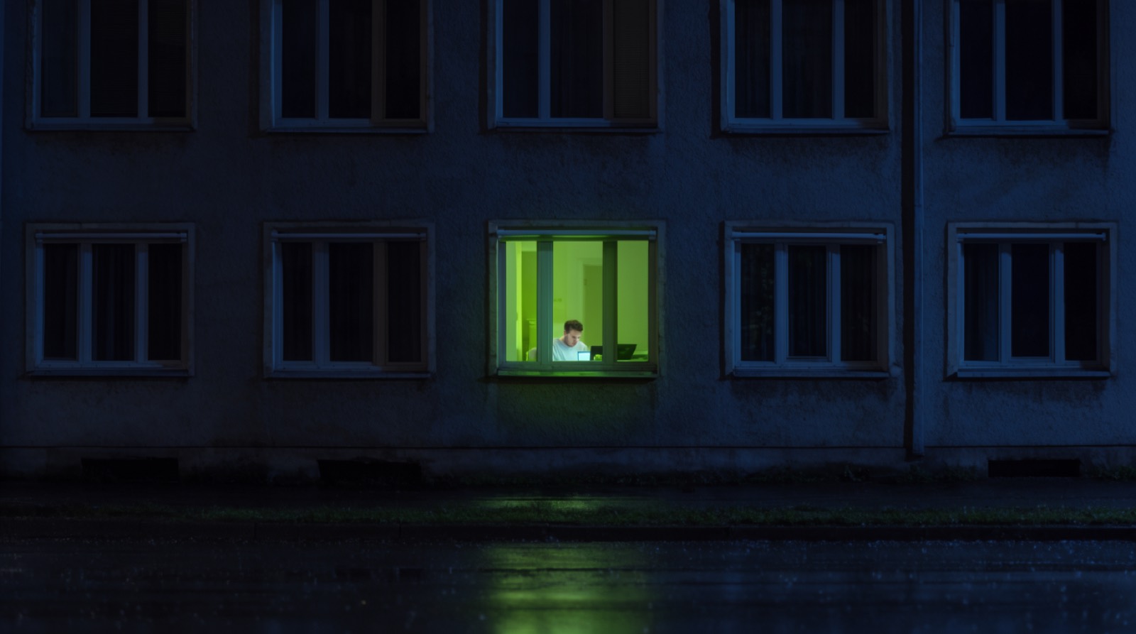 A dark apartment facade at night with one lit window, a man at a desk inside it, a lime glow from his screen on the window frame