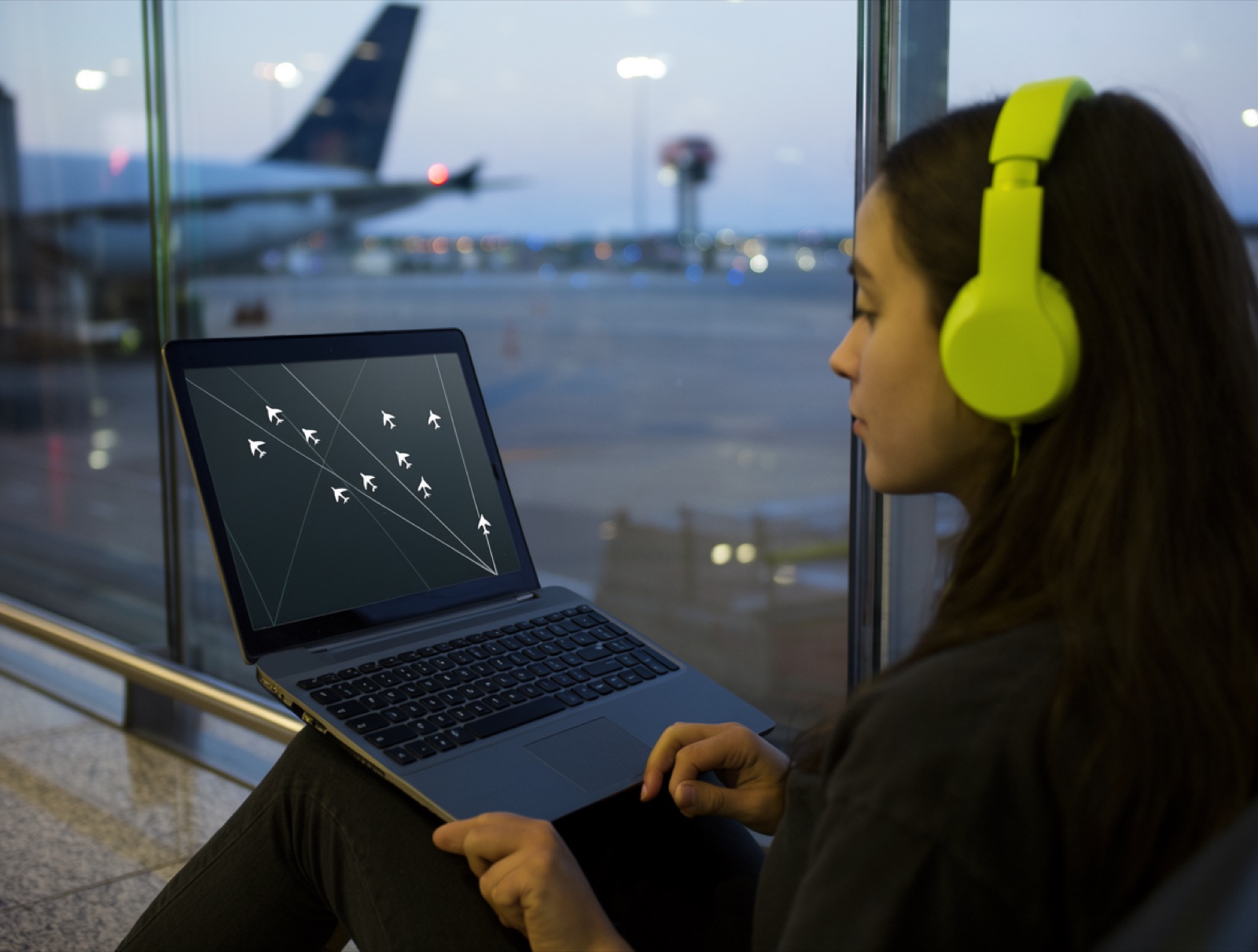 A young woman sitting against the glass wall of an airport gate at dusk, lime headphones on, a radar game on the laptop on her knees, an airliner blurred behind the glass