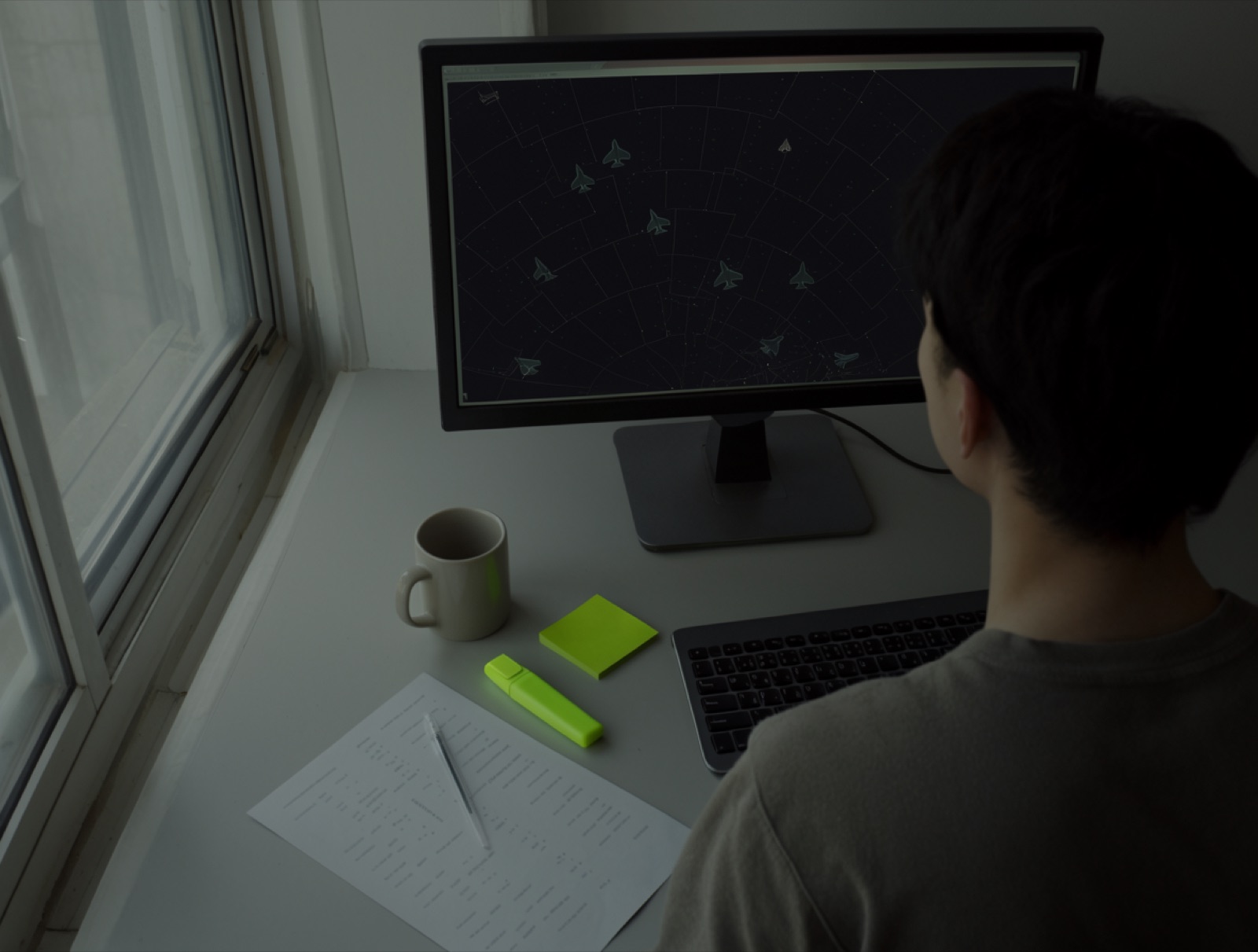 A student seen from behind at a window desk in morning light, an airspace diagram on a large monitor, a printed page, a mug, a lime highlighter