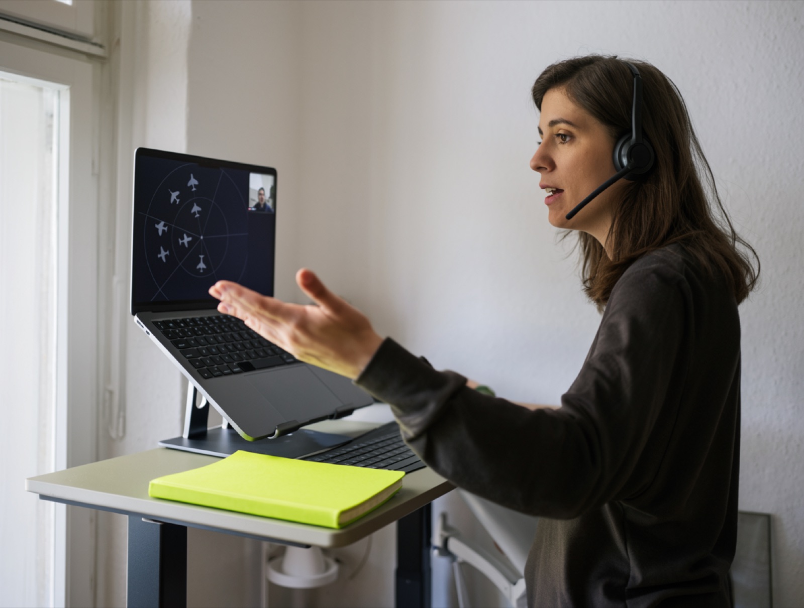 A young instructor in a headset standing at a desk in a bright room, gesturing towards a laptop that shows a radar training scene and a trainee in a small video window, a lime notebook on the desk
