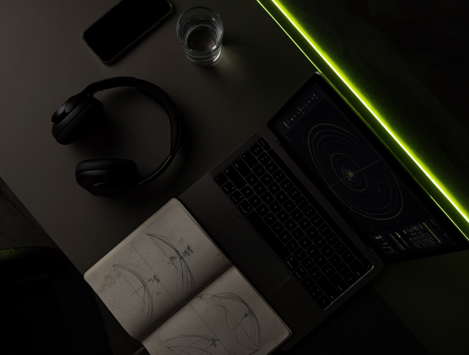 A desk seen from above at night: a laptop running a radar exercise, headphones, a notebook of sketched aircraft tracks, a glass of water, a lime light strip along the desk edge
